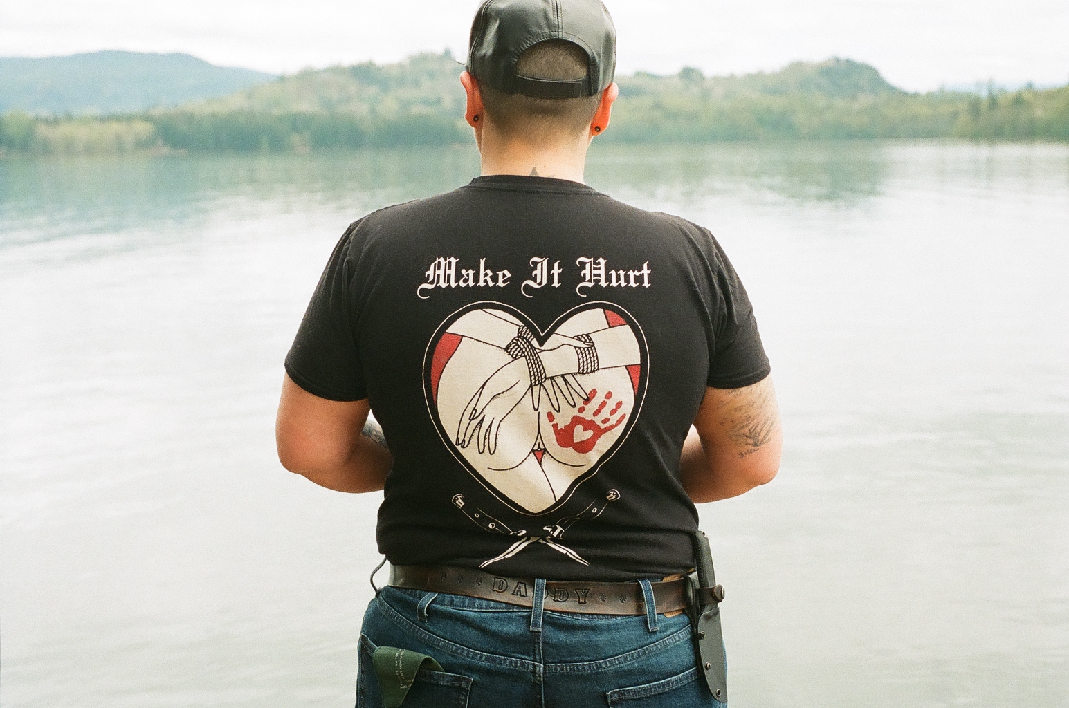 A butch standing in front of a lake with her back to the camera. Her shirt says make it hurt, and she has a hunter green hankerchief in her back left pocket 