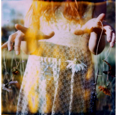 A girl wearing a dotted dress holds out her hands, each finger with a string attached to flowers. Her face cannot be seen.